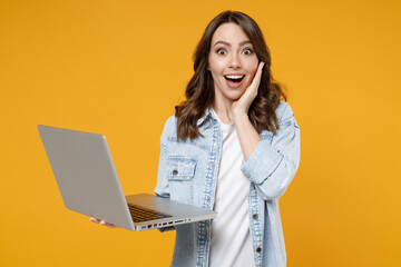 Young happy surprised shocked brunette fun woman 20s wearing stylish casual denim shirt white t-shirt hold laptop pc computer hold face look camera isolated on yellow color background studio portrait