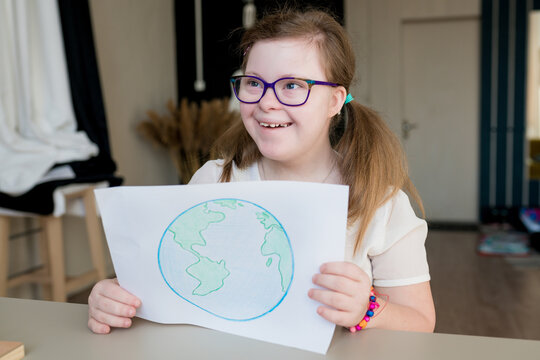 Portrait of teenage smiling girl with Down syndrome holding drawing of the planet Earth at home. Disabled child showing creative artwork. World Down syndrome day.