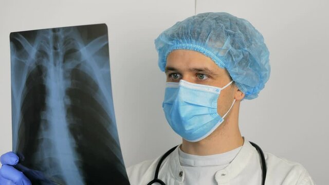 A young surgeon wearing a protective medical mask examines an X-ray of a patient's lungs and ponders the diagnosis.