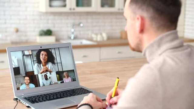 A smart guy is watching online webinar with a diverse people on the laptop screen, studying online, a man participates in virtual conference, taking notes, brainstorming with a workteam