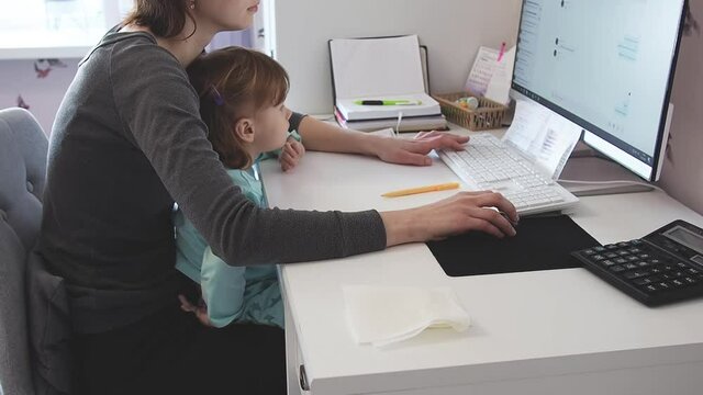 Mother multi-tasking, holding girl kid and using computer at home. Candid authentic and real life mom working and parenting
