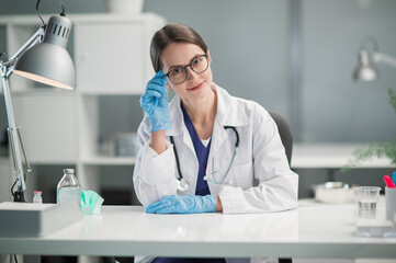 A woman doctor with glasses at her desk in the office is ready to receive patients of the clinic. A...