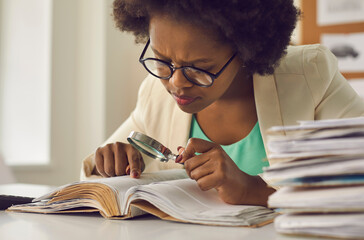 Closeup portrait young african american woman analyst expert with magnifying glass searching information in document sitting at desk with paper folder stack in comfort office. Research work
