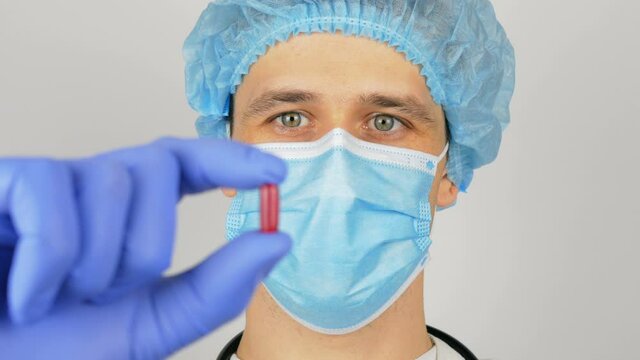 A handsome young doctor examines a new drug. A young handsome doctor in a medical mask holds a red pill in his hands presenting a new drug.