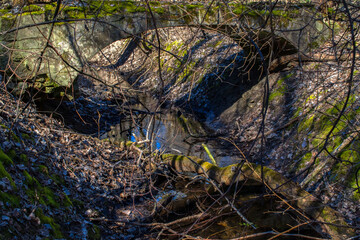 A stone bridge spanning the creek in the woods, with an outlined shadow on the shore