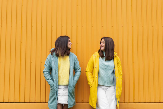 Sisters wearing yellow and blue raincoats smiling while looking at each other standing against wall