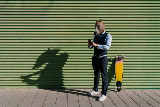 Male entrepreneur in horse mask using mobile phone while standing against wall