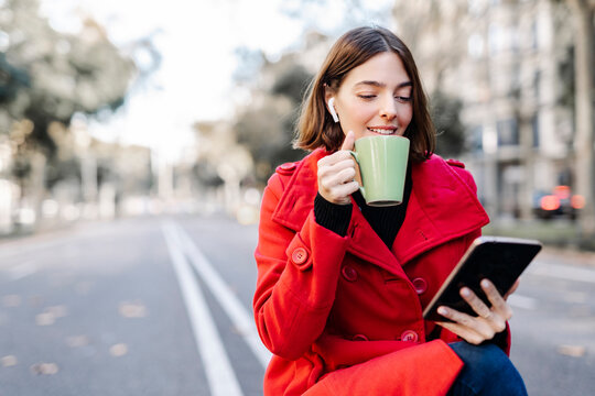 Smiling young woman in winter jacket having coffee while using digital tablet on road