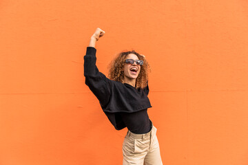 Cheerful woman with arms raised standing against orange wall