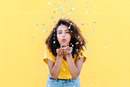 Young woman blowing multi colored confetti against yellow wall