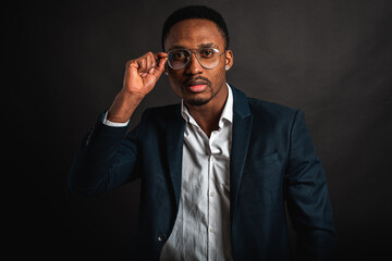 Portrait of attractive, handsome, stylish professional African American businessman in stylish suit and white shirt adjusts glasses by hand, isolated on dark background. Low key. Selective focus