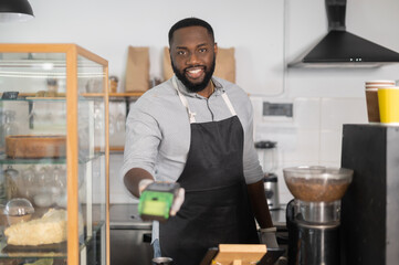 Cheerful and friendly multiracial waiter, African cafe manager, bakery staff is holding POS payment terminal and looks at the camera. Contactless payment concept