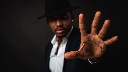 Young man in African American suit standing in front of camera with hand in front as stop sign, on dark background. Selective focus. Sign language.