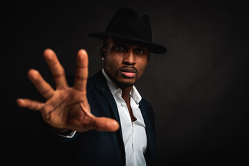 Young man in African American suit standing in front of camera with hand in front as stop sign, on dark background. Selective focus. Sign language.