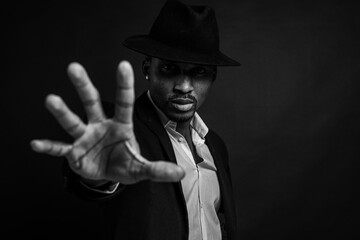 black and white photo, young man in African American suit standing in front of camera with hand in front as stop sign, on dark background. Selective focus. Sign language.
