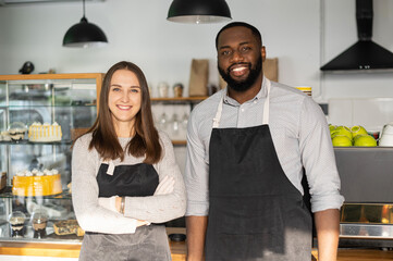 Smiling caucasian waitress and cheerful African-American waiter stands with arms crossed behind the counter. Confident multiracial baristas look at the camera