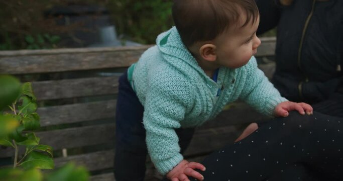 A little baby is sitting on a bench in the garden with his mother and his older sibling