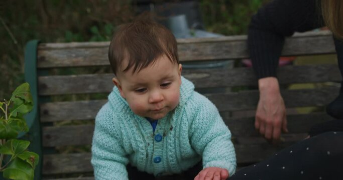 A little baby is sitting on a bench in the garden with his mother on a winter day