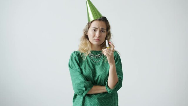 Portrait of miserable young woman wearing party hat blowing horn with sad and disappointed face standing on white background. People and bad celebrations concept.