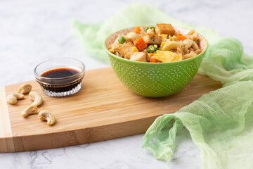 Bowl of Tofu Cashew Fried Rice with a Small Bowl of Soy Sauce and Cashews on a Board; Green Material in Background; White and Gray Marble Countertop