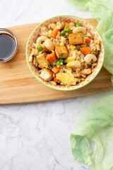 Top View of  Bowl of Tofu Cashew Fried Rice with a Small Bowl of Soy Sauce and Cashews on a Board; Green Material in Background; White and Gray Marble Countertop