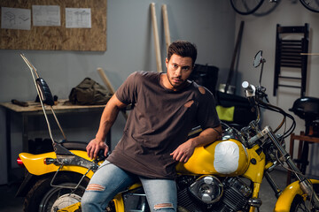 Fototapeta na wymiar A handsome young motorcyclist in jeans and a t-shirt poses for a photo sitting on a bike in his garage.