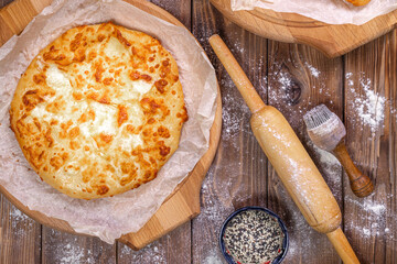 Khachapuri lies on wooden round pizza boards, chicken eggs and a rolling pin on a brown board background.