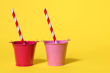 Two toy buckets on a yellow background with striped straws. The buckets are red and pink. The...