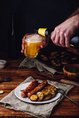 Pork Sausages Baked with Eggplant, Leek and Herbs on White Plate. Man Pouring Beer from Bottle into a Glass.