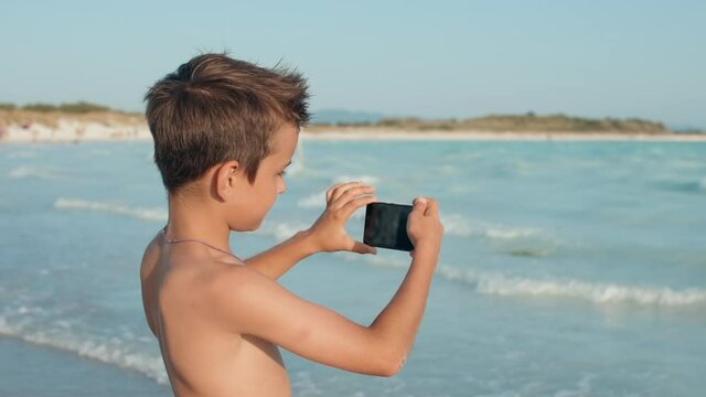 Joyful boy looking to screen at seaside. Teenager filming on camera at beach.