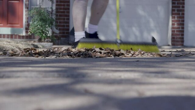 A senior adult homeowner sweeping up leaves in his driveway using a large push broom.