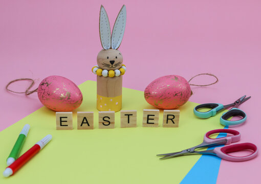 easter bunny and eggs.
Colored paper with scissors, eggs, bunny and the word Easter made from wooden cubes on a pink background, close-up side view.