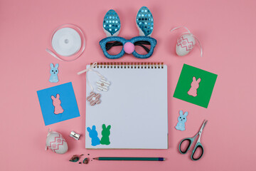 easter card with eggs and bunny.
Notepad, scissors, glasses, pencil, colored paper and a bunny on a pink background with a place for text in the middle, close-up top view.