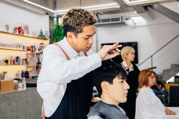 Asian male customer getting hair cut with hairdresser in salon. Fashionable barber using a scissor...