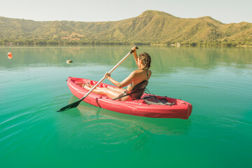 Latin woman paddling stand up on paddle, she is enjoying her vacation, she is in a red kayak on a turquoise lake and mountains in the background, behind her a summer sunset