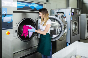 Female worker loads the laundry clothing into the washing machine at the dry cleaners. - obrazy, fototapety, plakaty