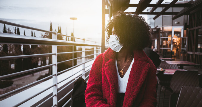 A portrait of a young curly African female in a virus protective mask and red demi-season coat sitting in an outdoor part of a roof street restaurant and pensively looking aside on a sunny evening