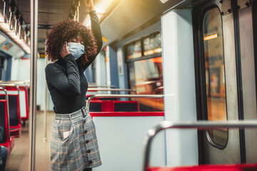 Portrait of a young curly African woman in a black turtleneck, plaid skirt, and a virus protective mask, standing and holding onto the handrail on a departing from a subway platform metro train