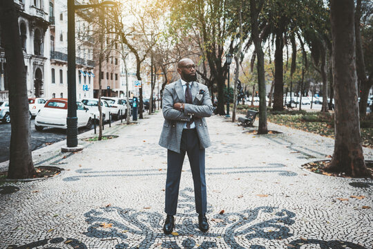 A stately mature hairless barbate black man entrepreneur in a dapper custom made suit is standing with hands crossed on the paving stone in the middle of a city alleyway surrounded by trees and cars