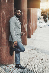 Vertical shot of an elegant bearded bald black businessman in a fashionable suit, standing on the street and holding and holding his eyeglasses in the hand leaning against a column of a bank building