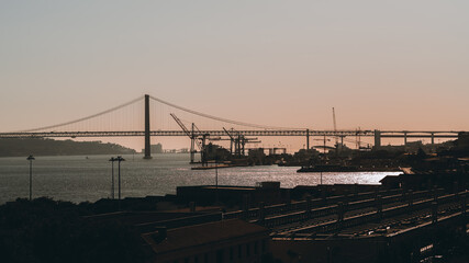 Sunset in Lisbon, Portugal: silhouettes of the Bridge 25 de Abril and docks in the background, roofs of houses, a train station in the foreground, strong specular on the water of Tagus river, pink sky