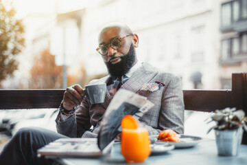 Portrait of a baldheaded adult black dandy guy with a well-groomed beard, in a custom made elegant costume and eyeglasses sitting in a street cafe with a cup of delicious coffee and reading a magazine