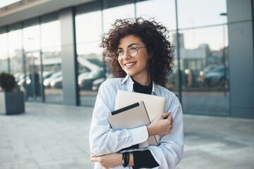 Curly haired business lady is embracing and holding a laptop while posing with glasses in front of...