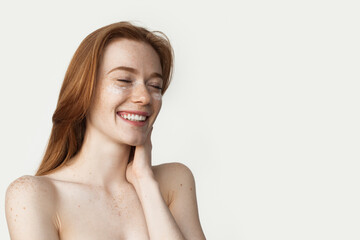Ginger woman with freckles is applying cream on her face smiling on a white wall with bare shoulders