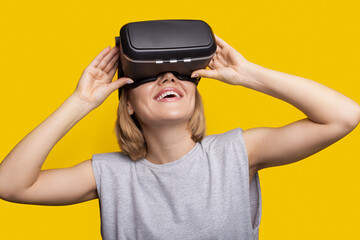Caucasian woman with blonde hair is smiling while testing new virtual reality headset on a yellow studio wall
