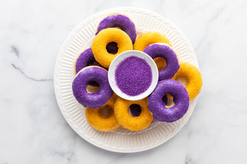 Top down view of a pile of purple and yellow iced donuts on a white plate resting on a marble table.