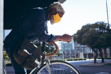 African american senior man wearing face mask leaning on bicycle in street using smartphone