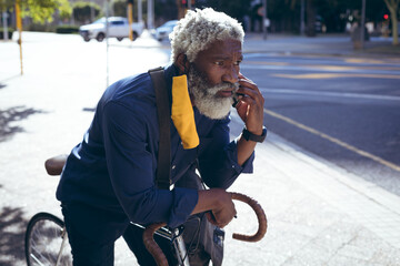 African american senior man wearing face mask leaning on bicycle in street talking on smartphone