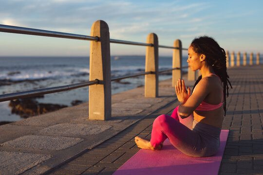 African american woman exercising on promenade by the sea doing yoga