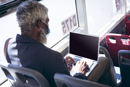 African american senior man wearing face mask sitting on bus using laptop